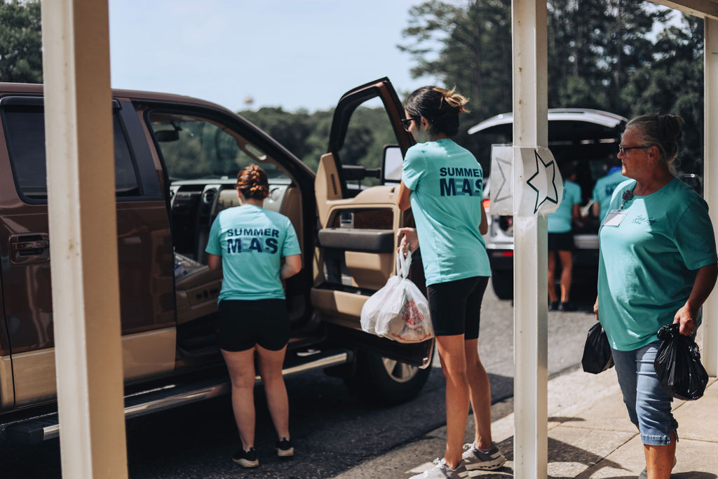 A few people in teal shirts, holding bags, stand by a car with its door open.