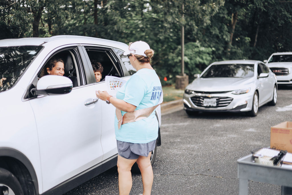 A person hands an item to a seated person in a car, while another person stands nearby.