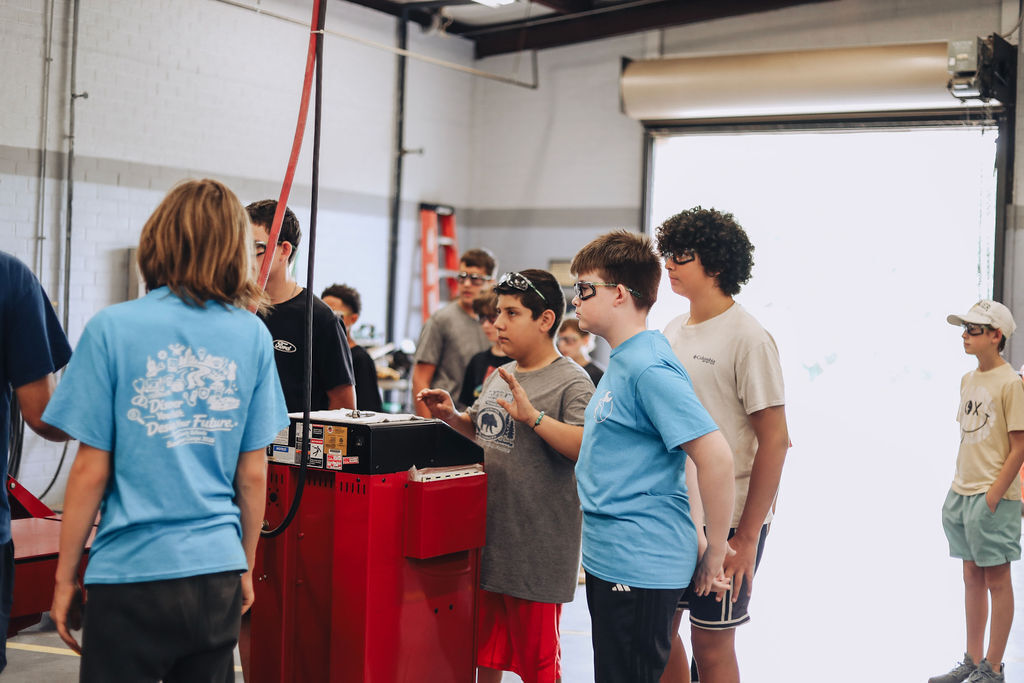 A group of kids in a workshop. One kid looks at a red machine while others listen.