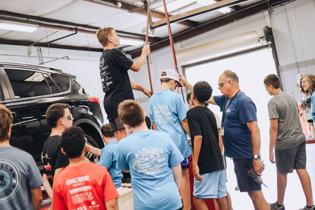 A group of people in a garage, some in light blue and black shirts, watching a person in a black shirt lift a car.