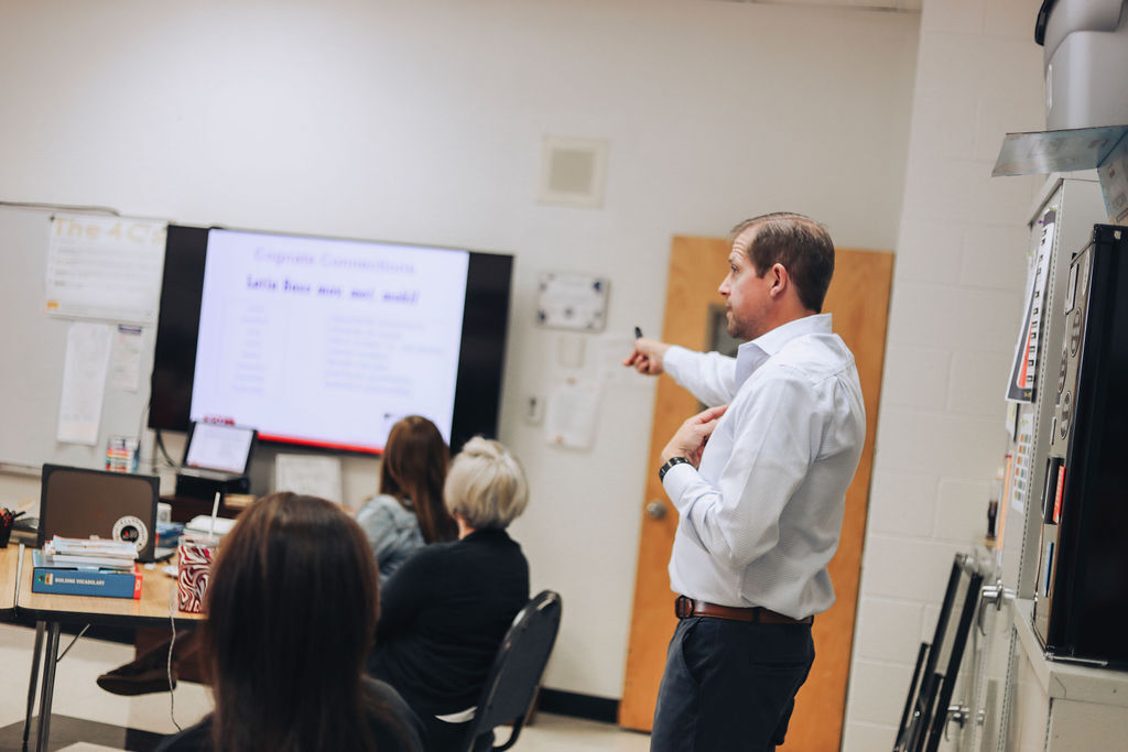 A man with a white shirt and brown belt speaks in front of a screen. Behind him are four people sitting in chairs.