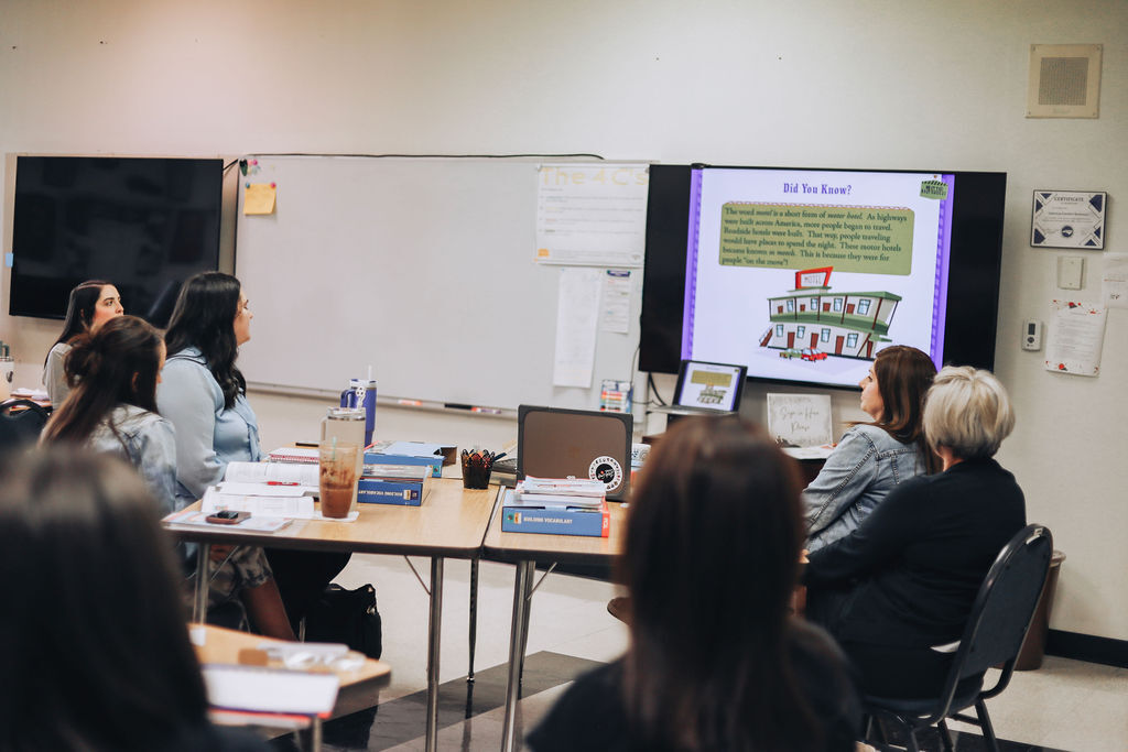People seated around a table listen to a presentation with a projector screen in the classroom.