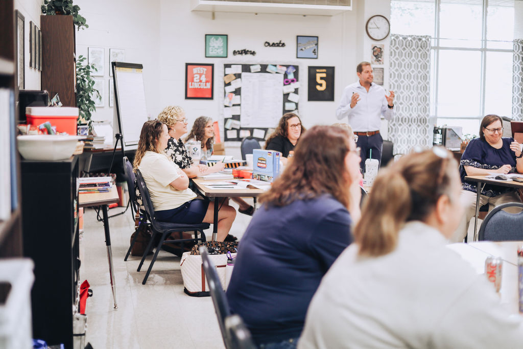 A group of adults sit around tables in a classroom, listening to a man in front of them.