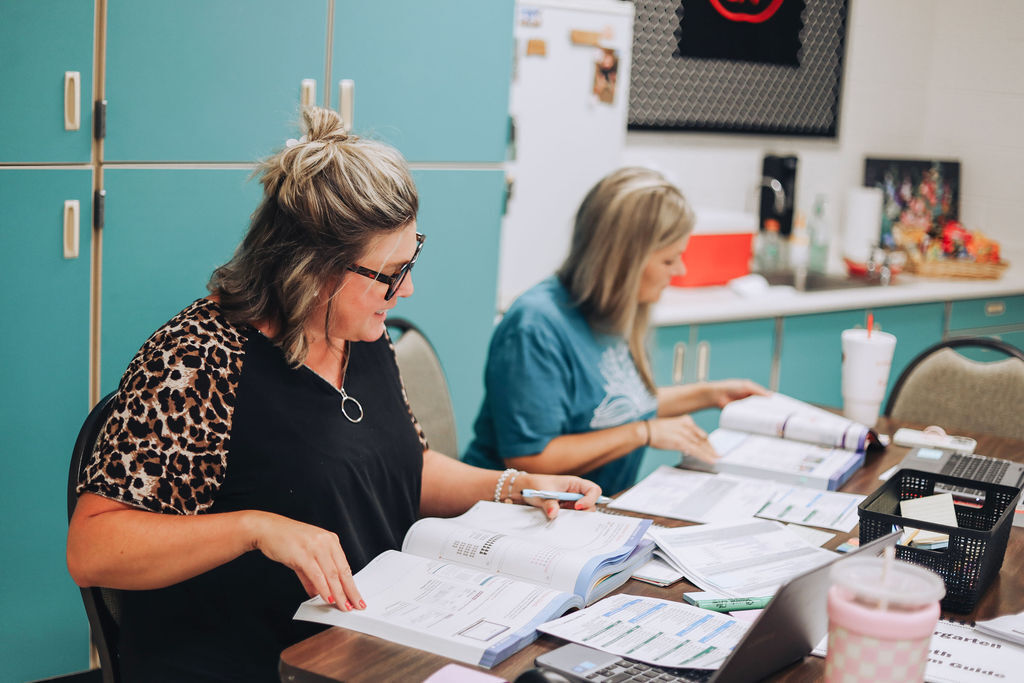 Two women at a desk, one wearing a leopard print blouse, the other in blue, both examining documents.