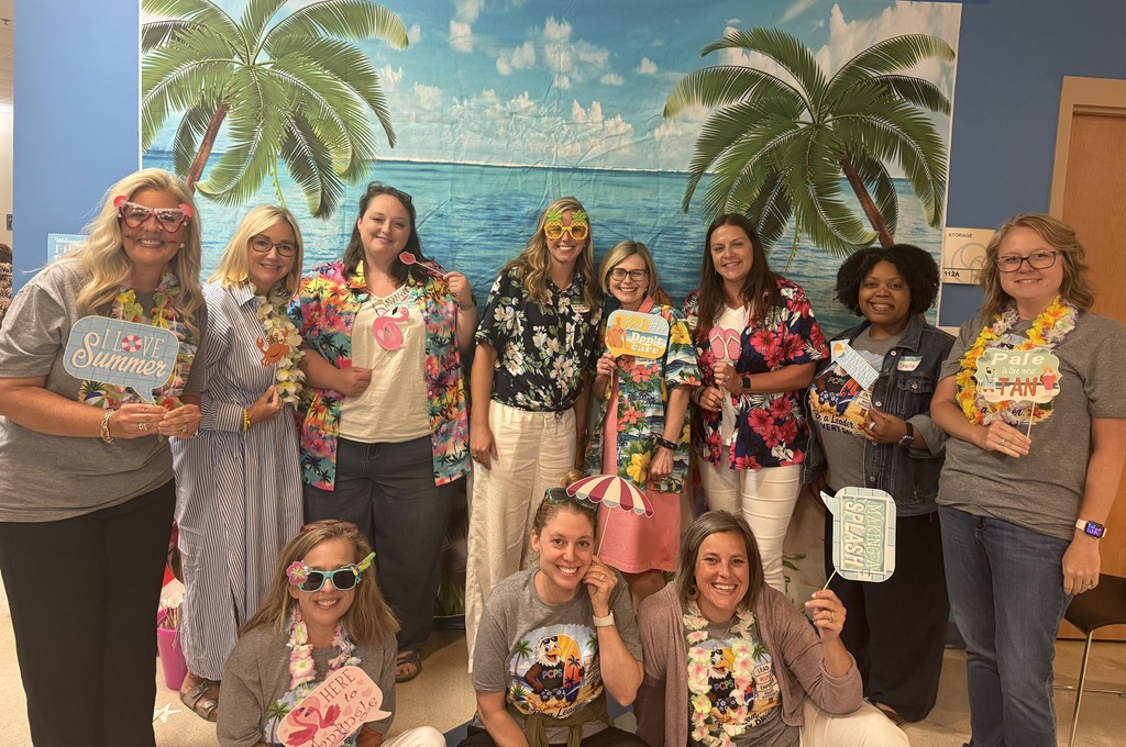 A group of women in tropical-themed shirts pose for a photo in a room. Background shows a mural.