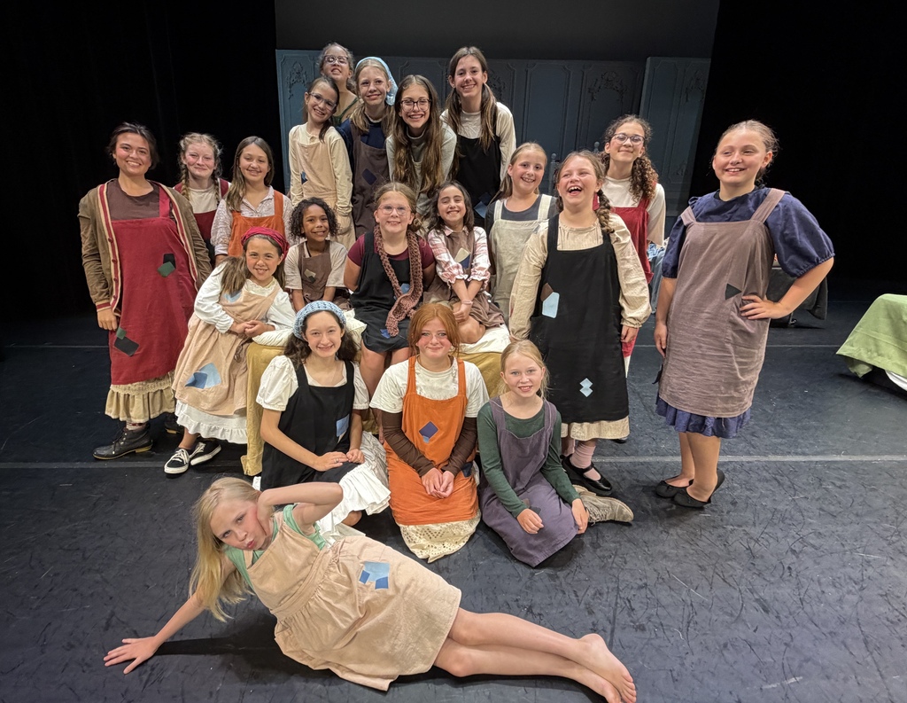 A group of young girls on stage, some kneeling or standing, wearing aprons and dresses, smiling for the camera.