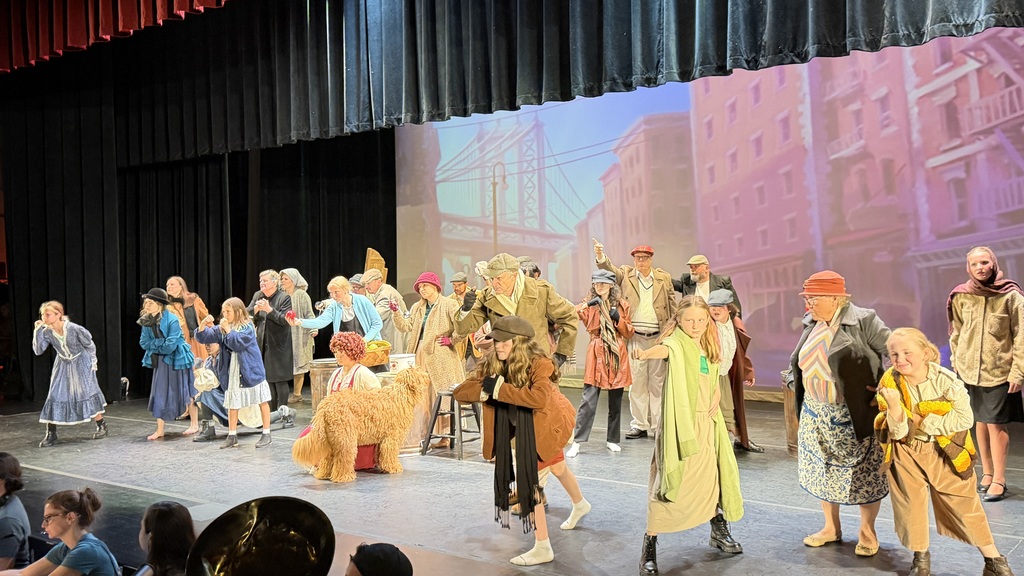 A group of people perform on a stage in front of an audience. Some wear hats.