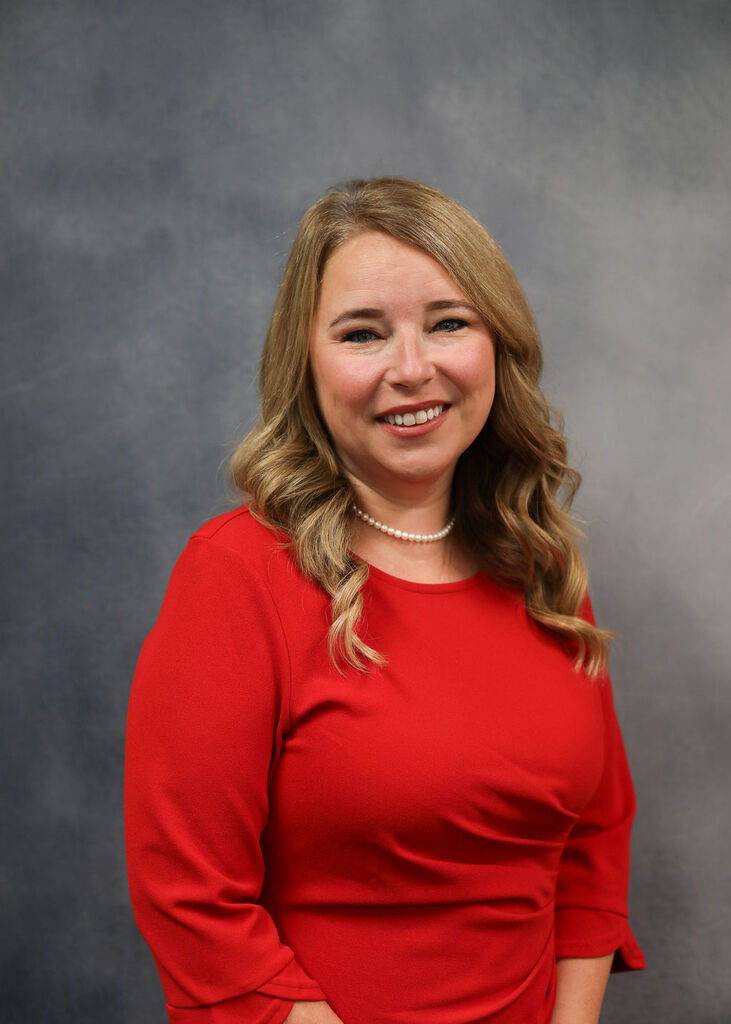 Woman with blonde hair, wearing a red blouse and pearl necklace, smiling and posing against a gray background.