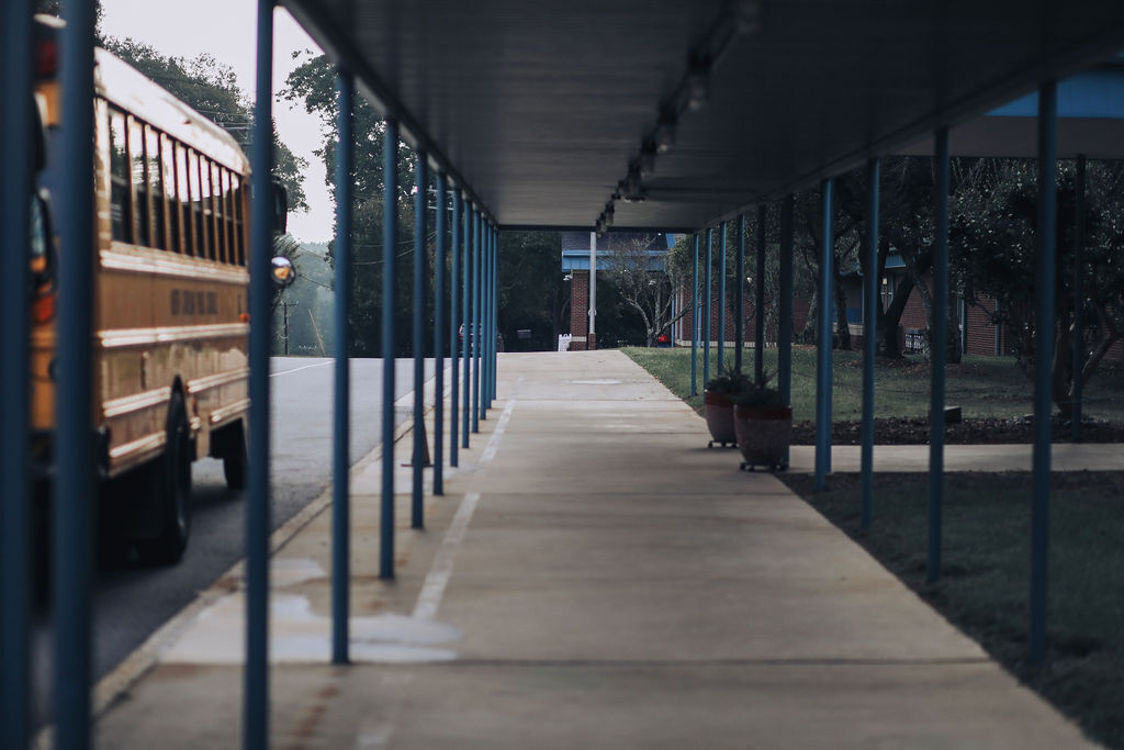 A school bus parked under a covered walkway, with blue poles on either side and a grassy area in the background.