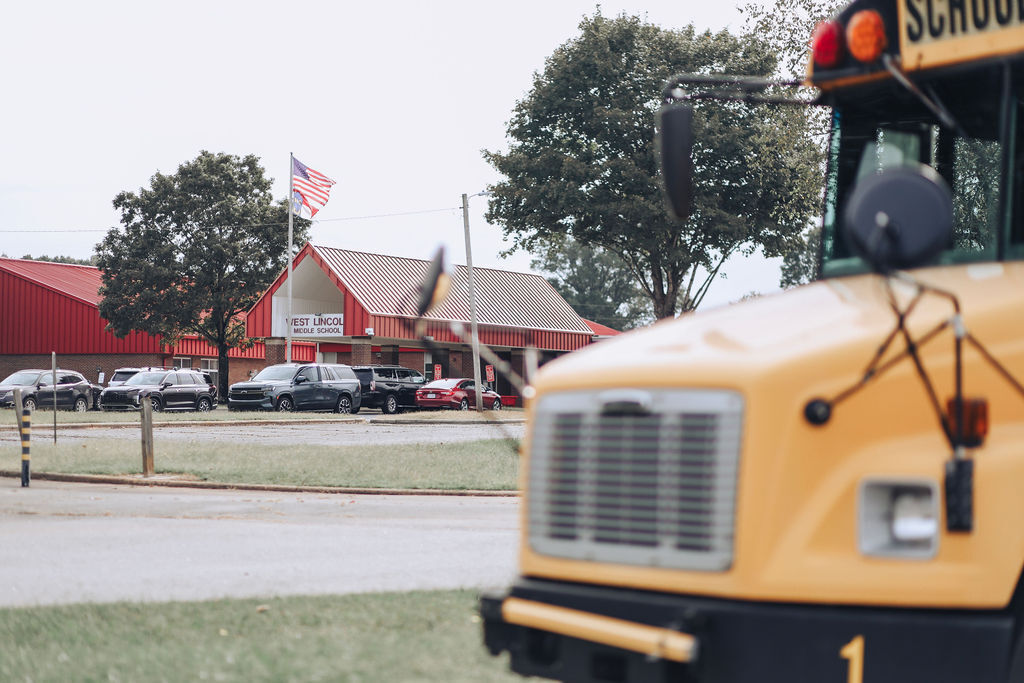 A yellow school bus in front of a building, with a flag and parked vehicles. Green grass and trees in the background.