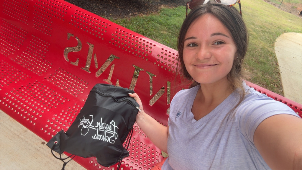 Woman sitting on a red bench, holding a black bag with white writing, wearing a gray shirt.