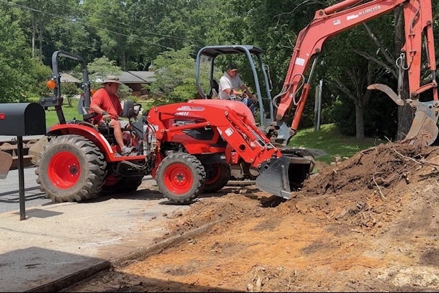 Two men are operating heavy machinery, one in a red tractor, another in a red excavator, working on a site.