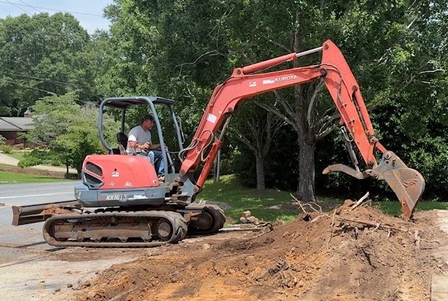 Red excavator parked on a road, operating on a pile of soil. Trees and a house in the background.