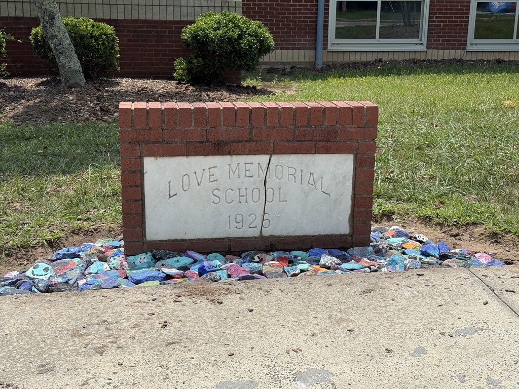 Brick sign with "Love Memorial School 1926" stands on sidewalk with colorful glass pieces. Behind, a building with windows and shrubs.