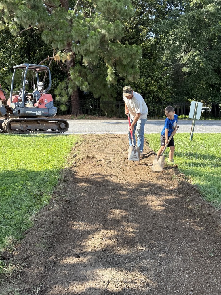 Two people work outdoors, one adult and a child. They use shovels on a dirt patch. Behind them, a large excavator operates. Trees and grass surround the area.