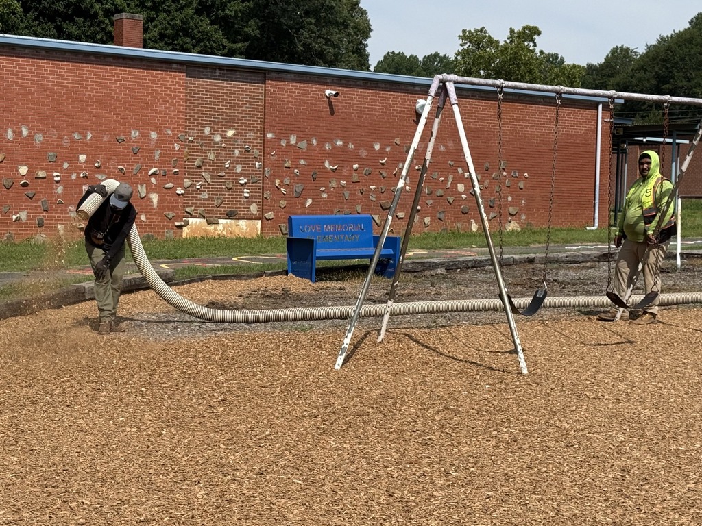 Two workers in yellow coats and masks using hoses to clean a playground. A brick building with a textured wall is in the background.