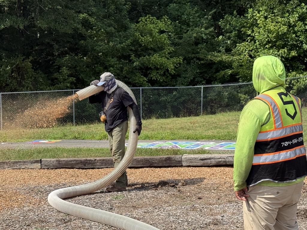 Two workers in safety gear pour sand from a hose onto a gravel area. A fence and trees in the background.