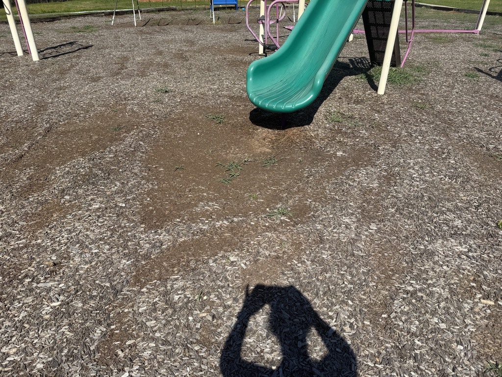 A playground with a green slide, a shadow of a person, and a gravel surface.