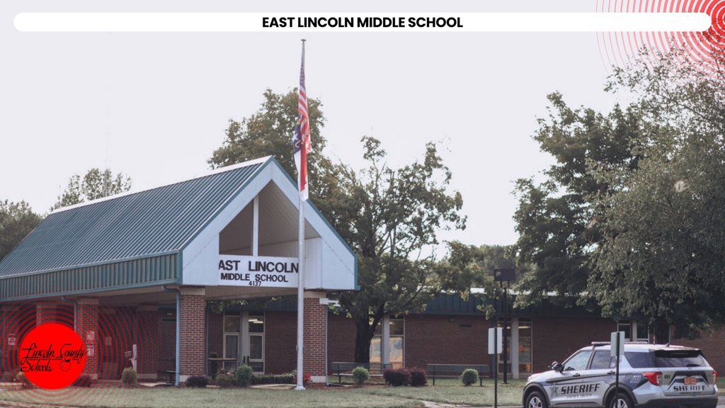 A school building with a gray roof and red trim stands in front of trees. A flagpole is nearby.