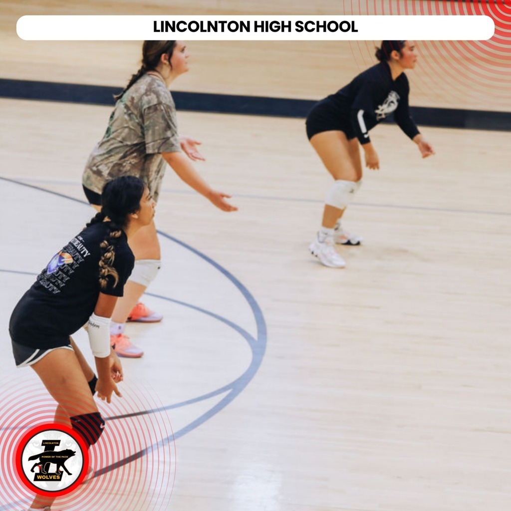 Three women play volleyball on a wooden court. Two are in action, and one watches. They wear matching black jerseys.