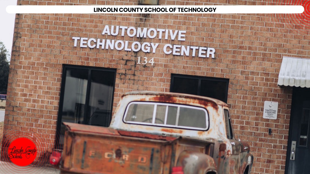 A rusty pickup truck parked in front of the Automotive Technology Center, a brick building with a sign.