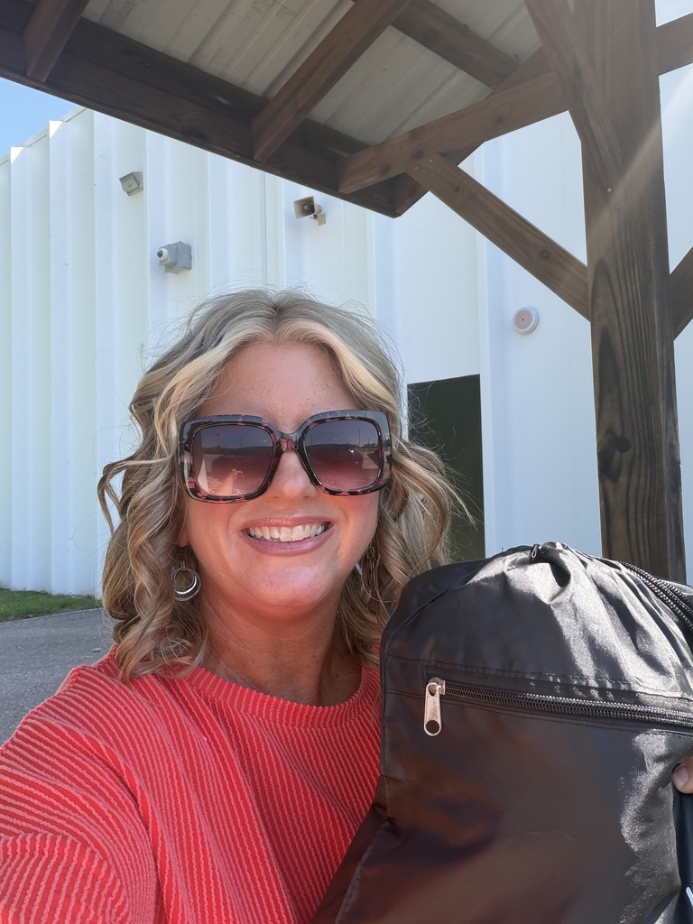A woman with blonde hair wearing sunglasses and a red shirt holds a black backpack. She is smiling. Behind her is a white building with a wooden roof.