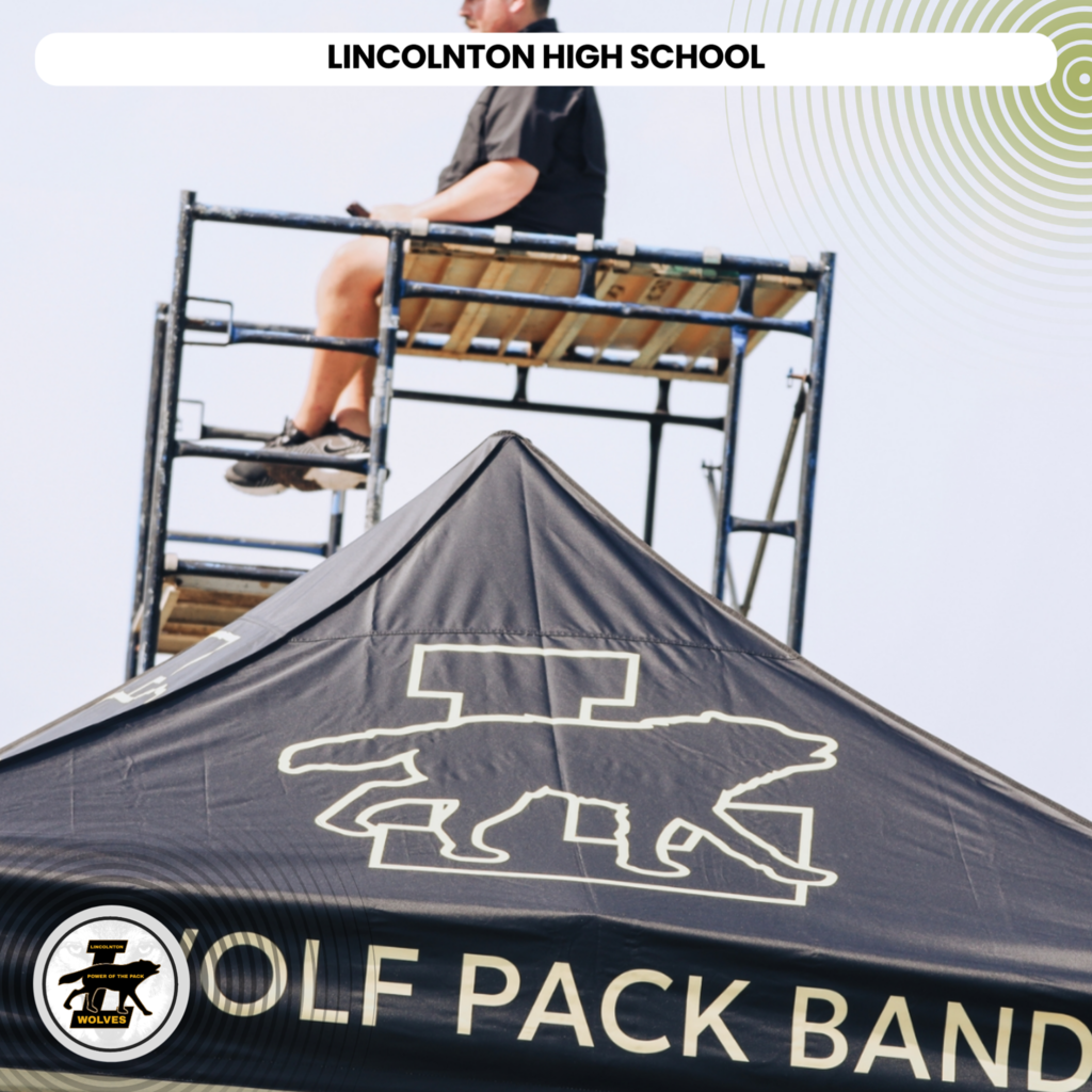 A man sits on a metal scaffold above a tent with a wolf logo. The tent reads "WOLF PACK BAND".
