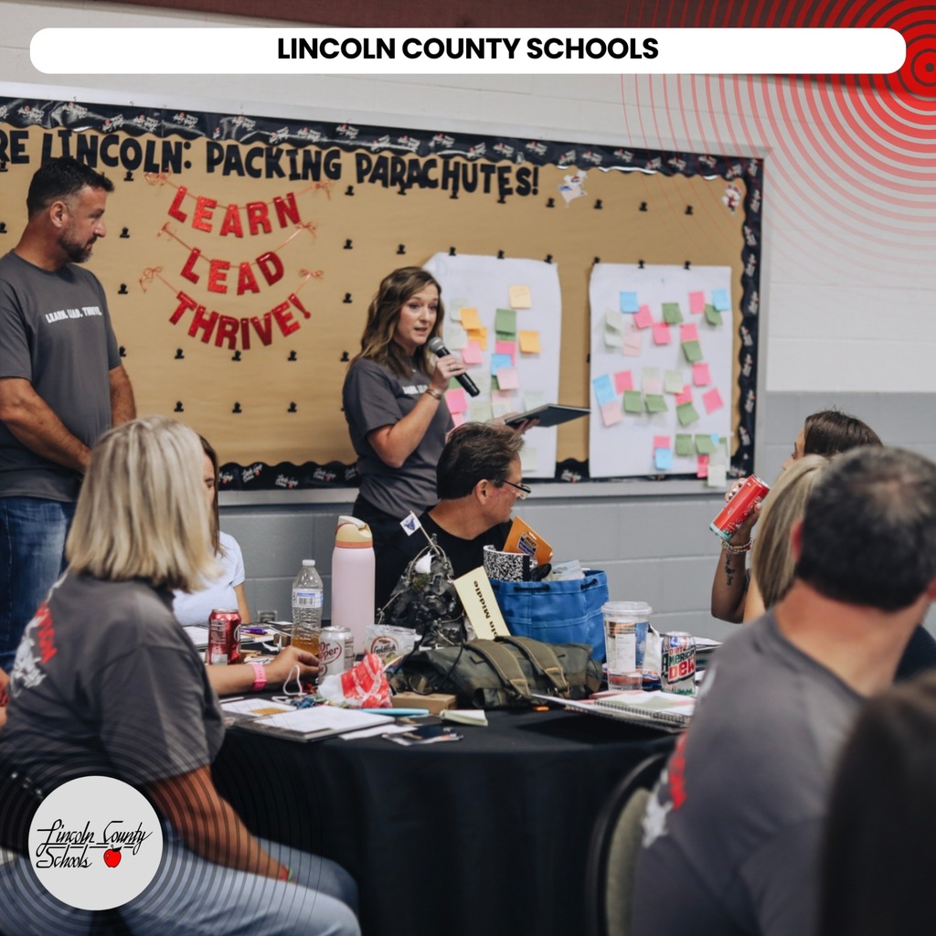 A group of people are seated at a table in a classroom. A woman is speaking into a microphone in front of a bulletin board.
