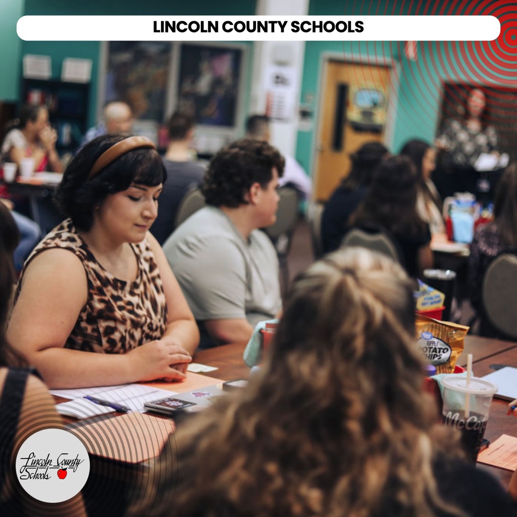 A woman sits at a table with others, all facing forward. A sign at top reads "Lincoln County Schools".