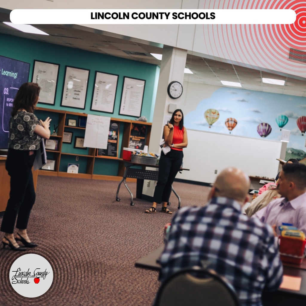A group of people in a classroom. A woman in red stands by a table. A clock and balloons are on the wall.