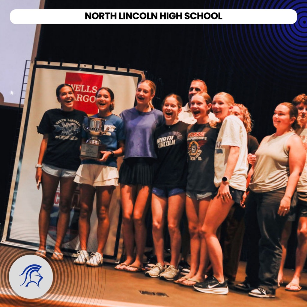 A group of students stand together on a stage, with one holding a trophy. They wear sports attire.