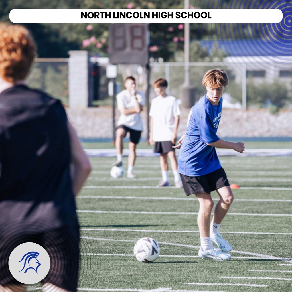 Four boys on a soccer field. One is kicking a ball and the others are watching.