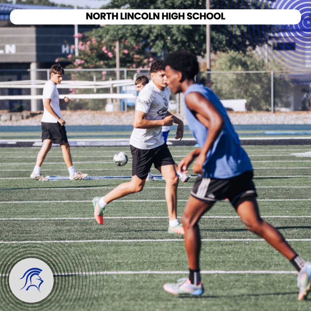 Four individuals play soccer on a field with a fence. The player in front wears a blue jersey.