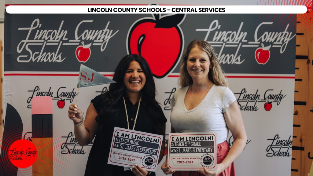 Two women stand near a backdrop. One holds a sign reading "I am Lincoln". The other displays a flag.