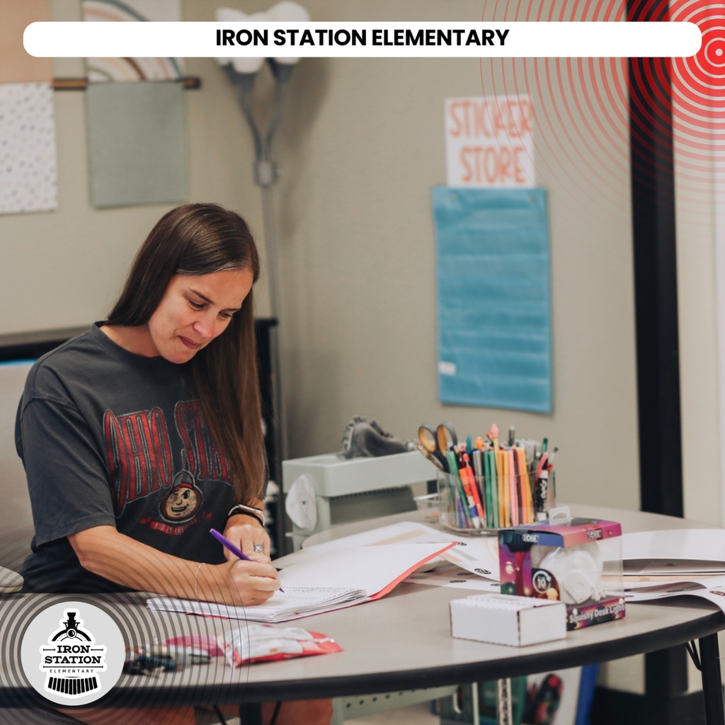 A person in a black t-shirt writes on paper at a table in a classroom with various school supplies.