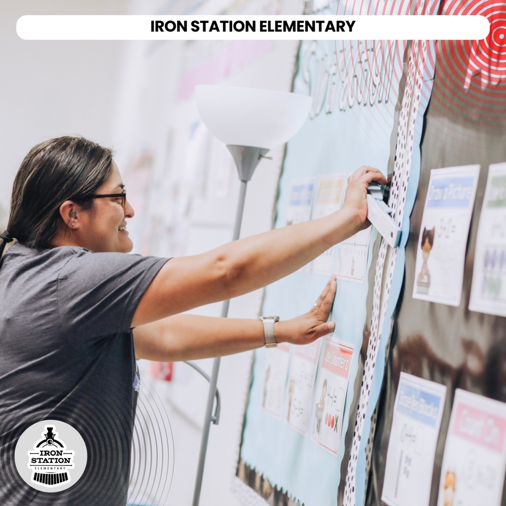 A woman in a gray shirt and glasses pins a paper on a bulletin board.