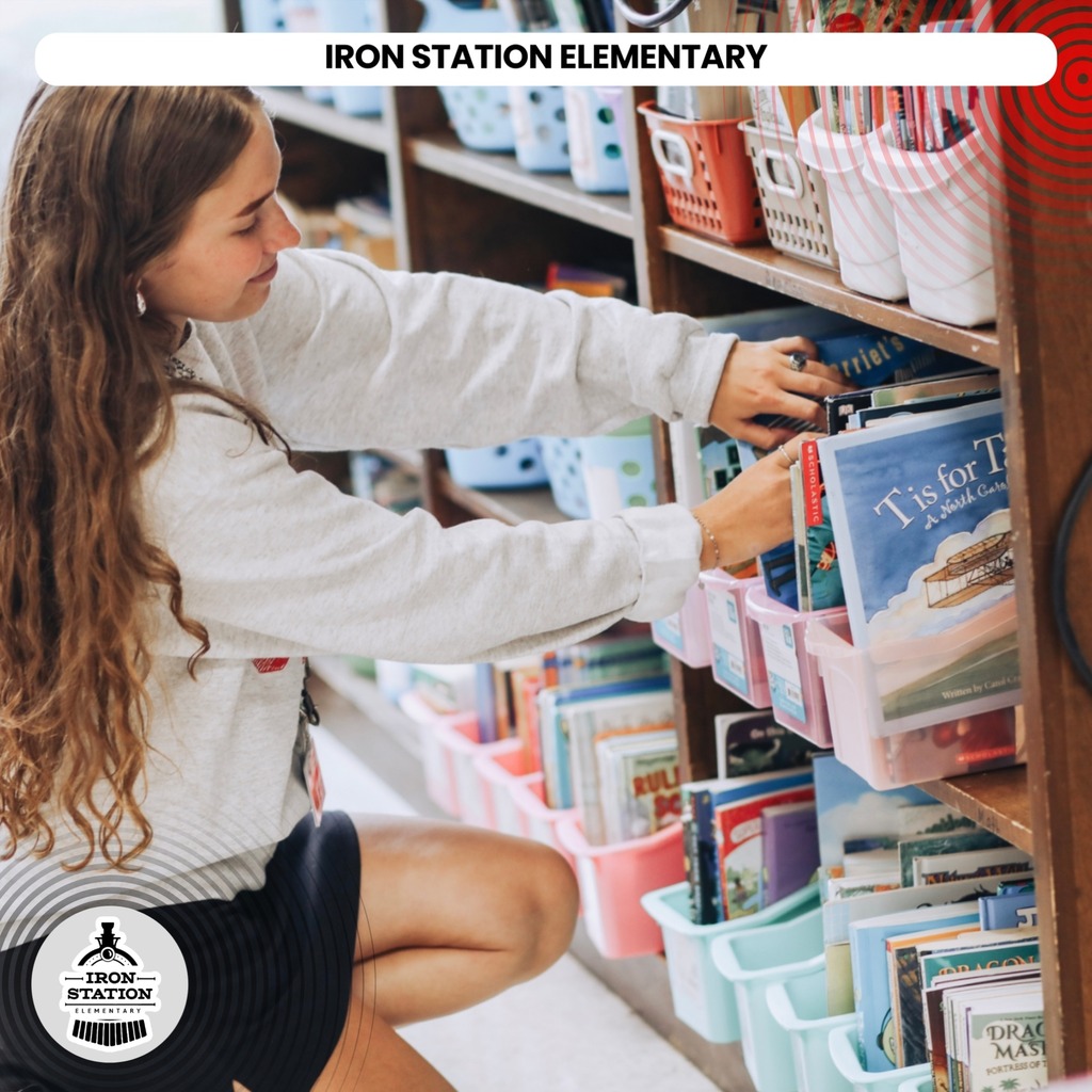 A girl with long curly hair kneeling, selecting books from a library shelf. The shelf has colorful storage bins.