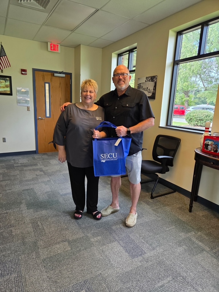 A man and woman stand in a room, smiling, with the man holding a blue bag. A window and flag are in the background.