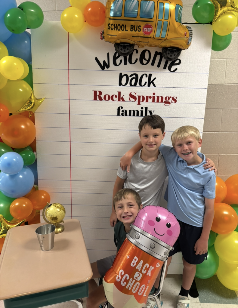 Three boys stand in front of a welcome backdrop, balloons, and a balloon bus. A large pencil balloon stands between two boys.