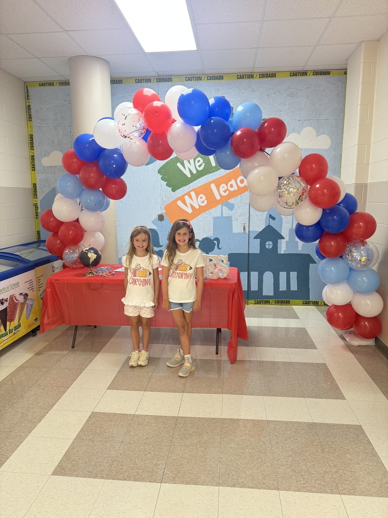 Two children stand under a balloon arch that reads "We Lead." A red table is in front of them.
