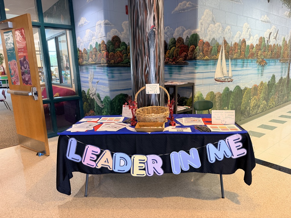 A table with a blue cover displays items including cards and a basket. "LEADER IN ME" is written across the front.