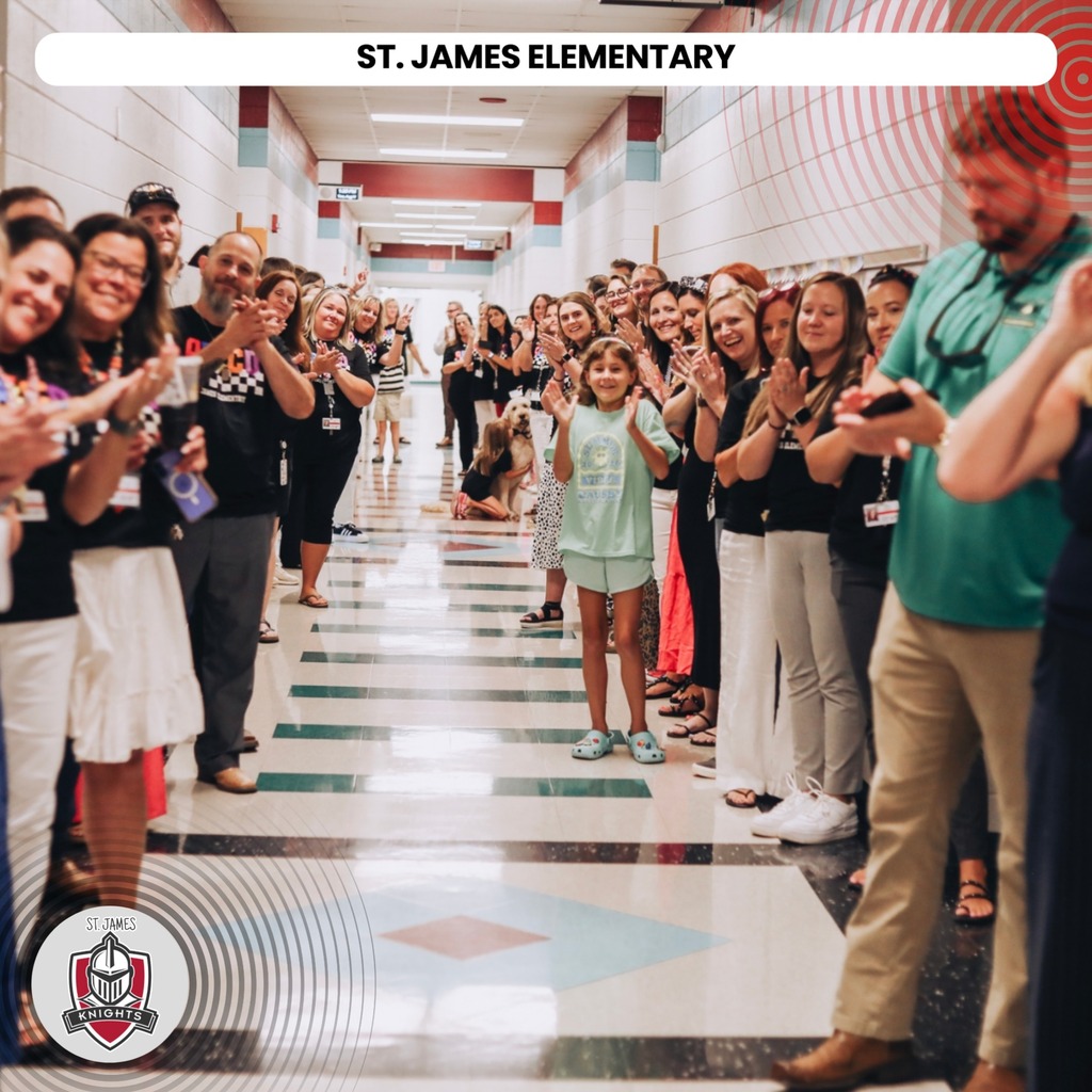 Many people stand in a hallway, clapping and smiling. They face a young girl in the center. The hallway has white tile flooring and text reading "St. James Elementary" at the top.