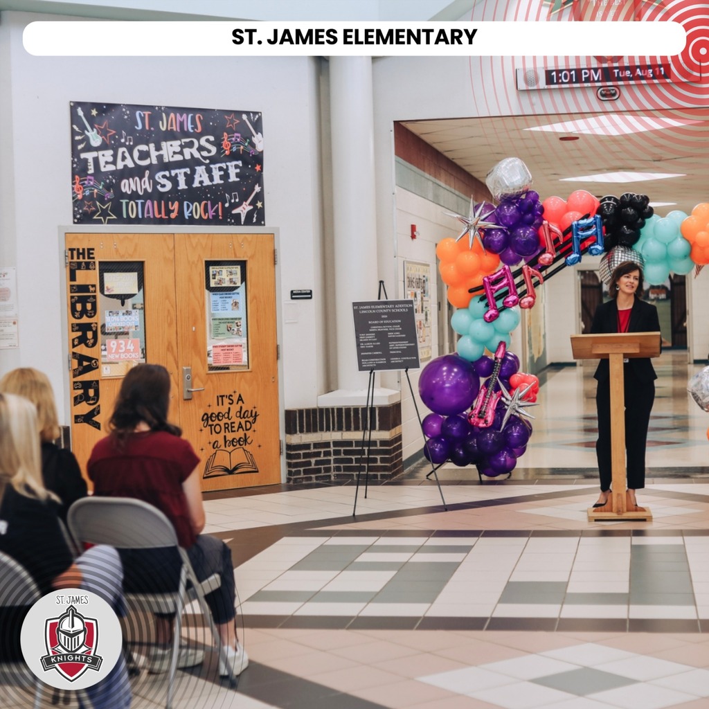 A woman stands at a podium in a hallway. Colorful balloons are behind her. A banner says "Teachers and Staff".