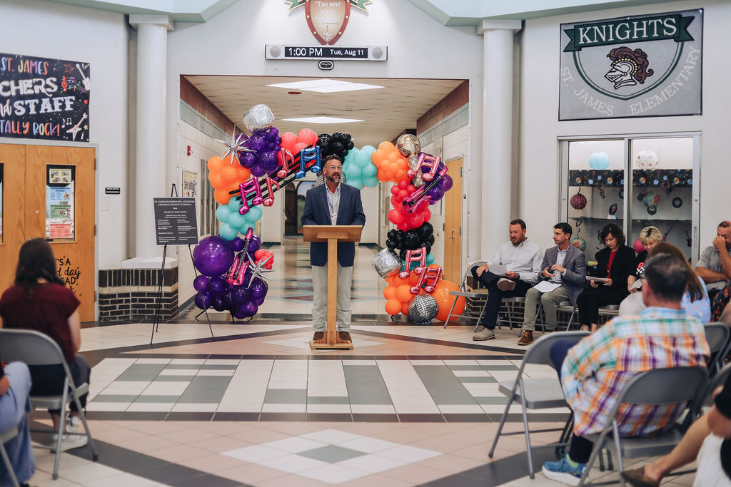 A man stands at a podium with balloons behind him, addressing an audience seated in a tiled room.