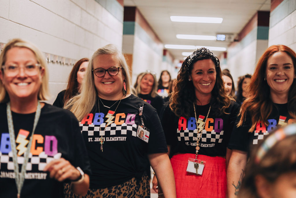 Several women wear black ABCO T-shirts, standing in a hallway, smiling at the camera. Background shows a long, well-lit corridor.