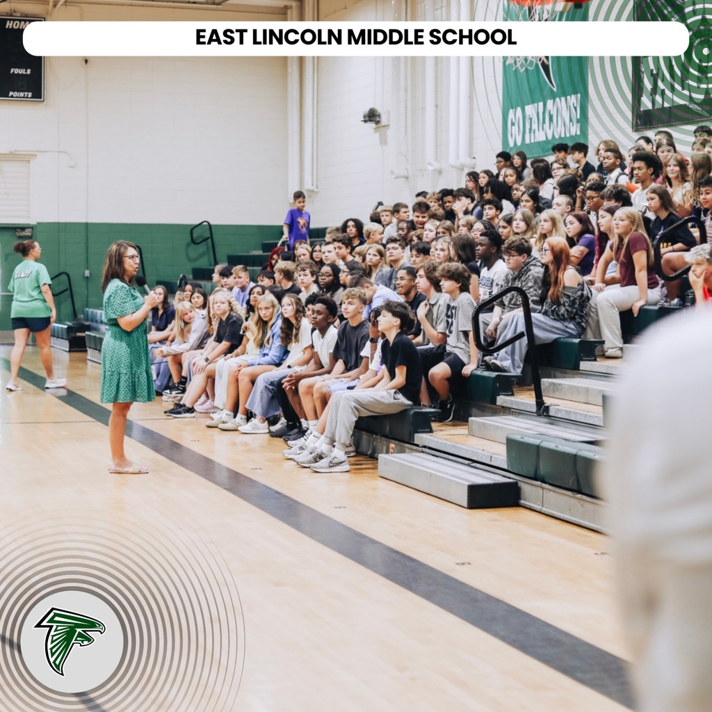 A crowd of students sit on bleachers in a gymnasium, with a woman in green speaking at the front.