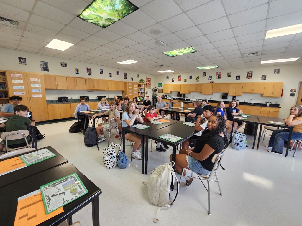A classroom with students seated at tables, bags and books around them, wooden cabinets, and a ceiling with lights.