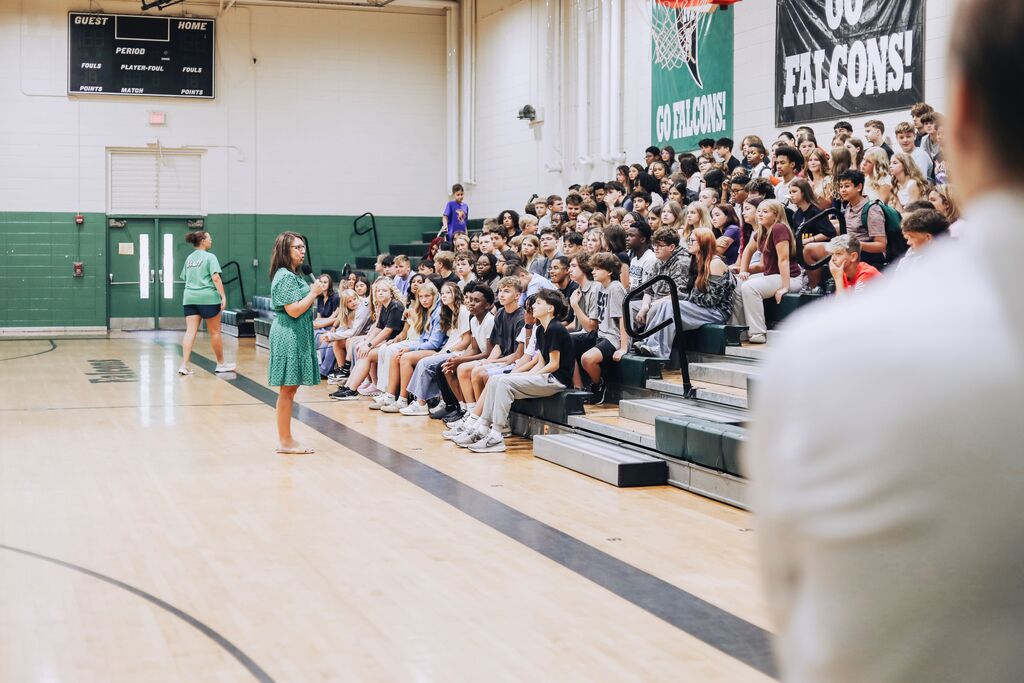 An indoor basketball court with people seated in bleachers. A woman in green stands in front of the crowd.