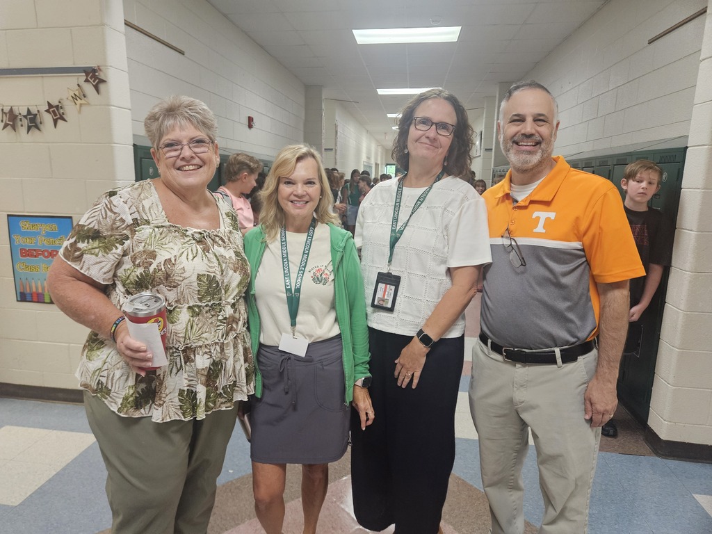 Four people standing in a school hallway with white walls, smiling and posing for a photo.