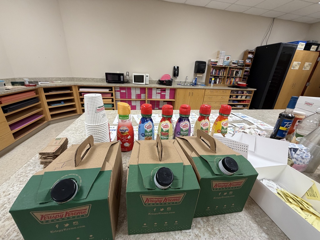 Green boxes with black lids sit on a white table, with several bottles and other items. A kitchen area is behind the table.